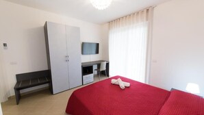 Ein Schlafzimmer mit roter Bettdecke, einem weißen Kleiderschrank, einem Schreibtisch mit Stuhl und einem an der Wand montierten Fernseher.