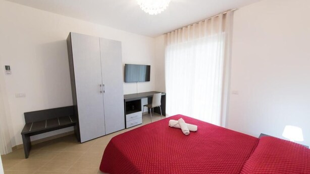 Ein Schlafzimmer mit roter Bettdecke, einem weißen Kleiderschrank, einem Schreibtisch mit Stuhl und einem an der Wand montierten Fernseher.