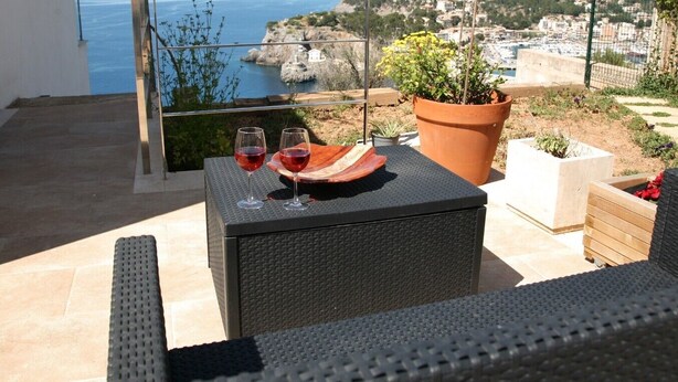 Un balcon avec vue sur la mer et la ville. Sur une table, on peut voir deux verres de vin rouge, une plante en pot et une boîte en bois.