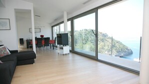 Un salon moderne doté d’une grande porte-fenêtre coulissante donnant sur un balcon offrant une vue panoramique sur la mer et les collines.