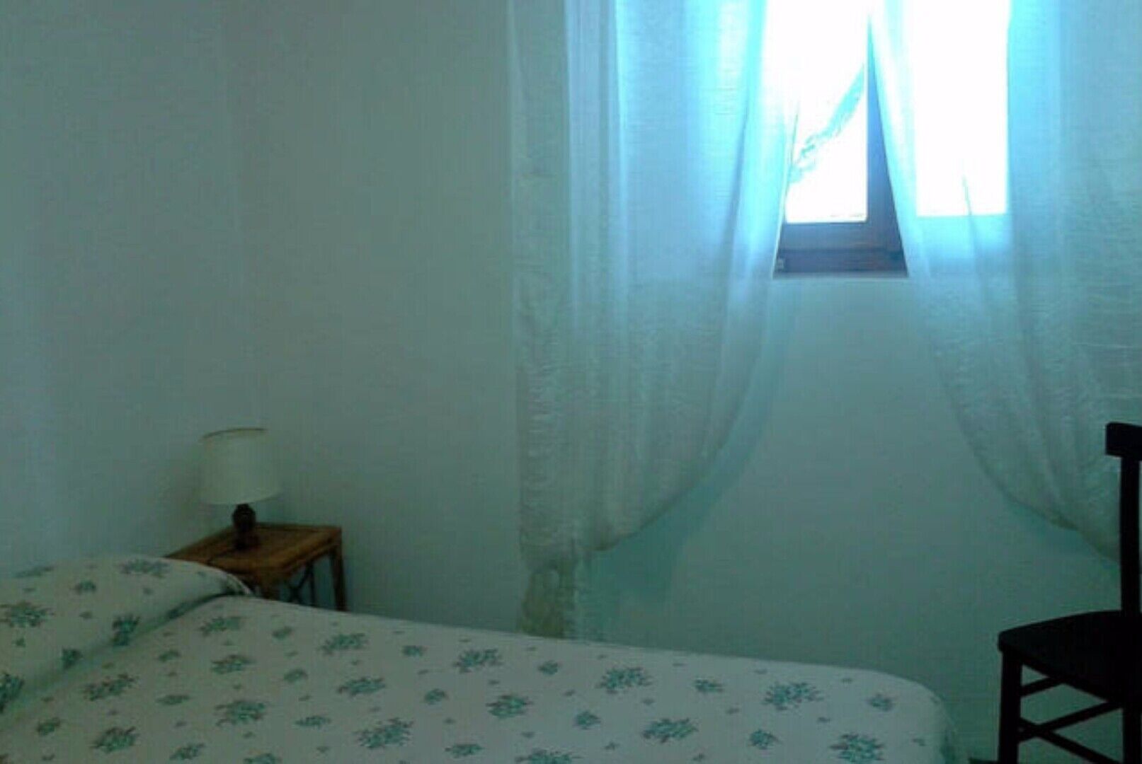 Ein kleiner, schlicht eingerichteter Raum mit einem Bett, einem Nachttisch und einem Stuhl.