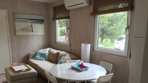 Un salon avec un canapé beige, une table ronde blanche entourée de chaises, un tableau accroché au mur et une fenêtre donnant sur des arbres et une voiture garée à l’extérieur.