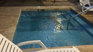 Une piscine avec un escalier en métal, entourée d’une terrasse en béton. On y trouve des chaises longues blanches et un espace de détente couvert avec une table et des chaises.