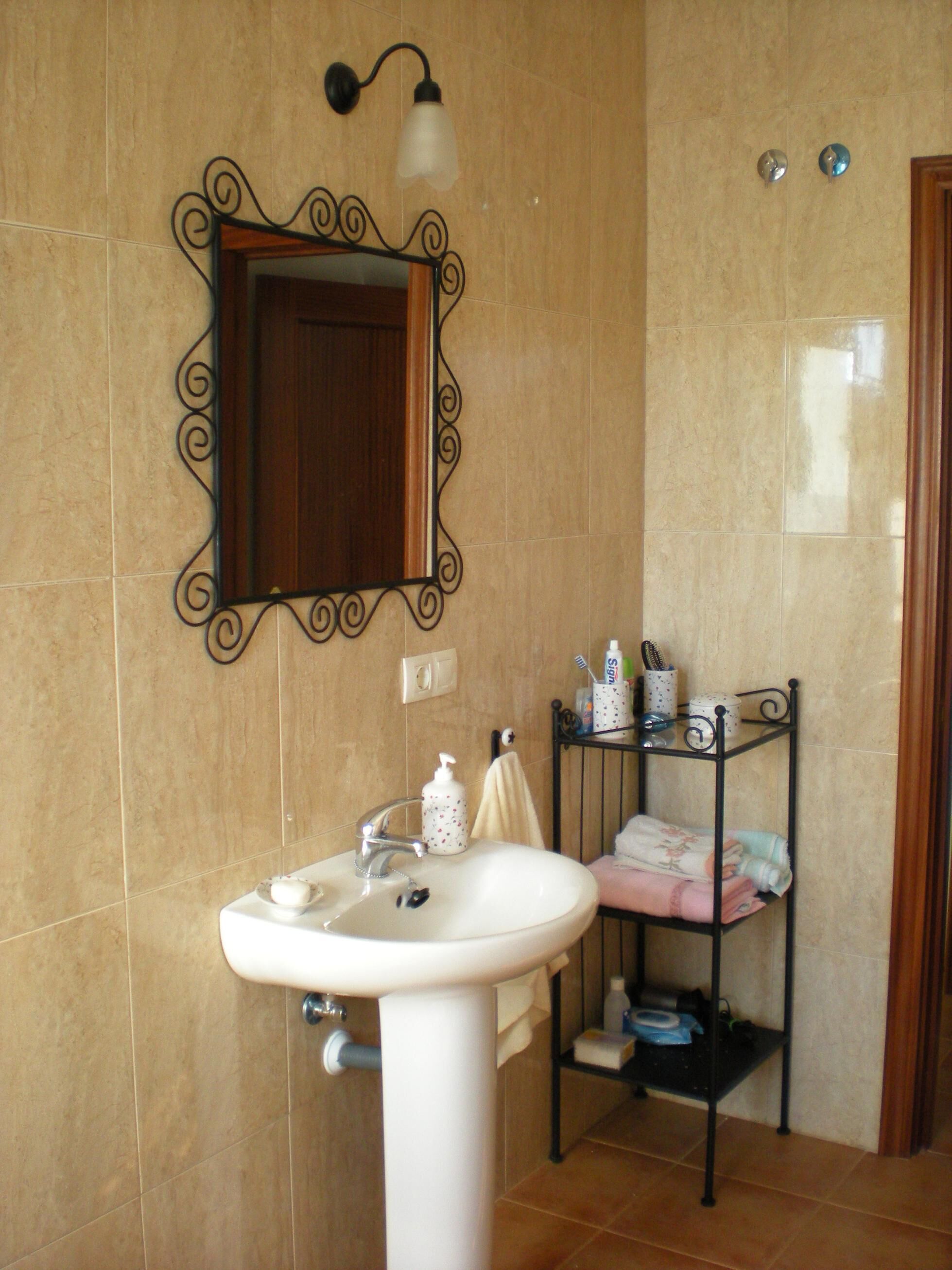 A bathroom with a white sink, a mirror with a decorative frame, a wall-mounted light fixture, and a shelf with toiletries.