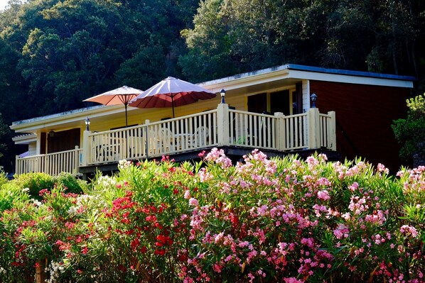 A two-story building with a balcony, surrounded by lush greenery and flowering plants.