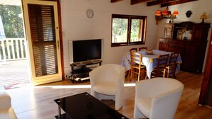 Ein Wohnzimmer mit Fernseher, weißen Sesseln, einem Esstisch mit floralem Tischläufer und einem Holzschrank.