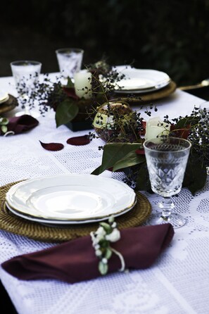 A beautifully set table with a lace tablecloth, greenery, and white flowers, featuring fine china, glassware, and a candle.