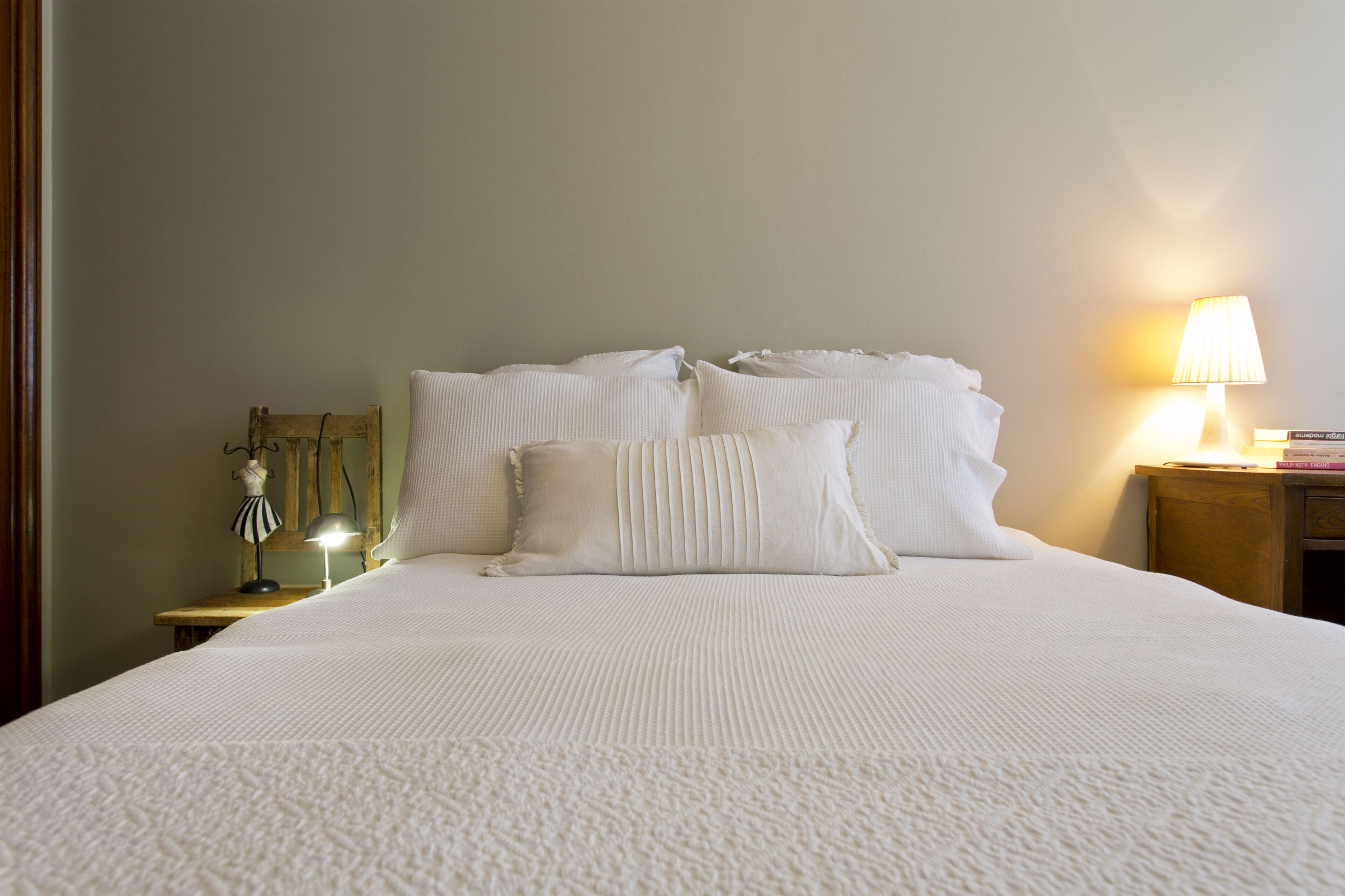 Un lit bien fait, avec du linge de lit blanc et des oreillers, une lampe de chevet et une petite table de nuit en bois sur laquelle se trouvent une lampe et un objet décoratif.
