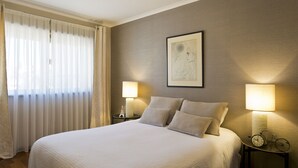 Una cama bien hecha con edredón blanco, dos lámparas de noche y una obra de arte enmarcada en la pared.