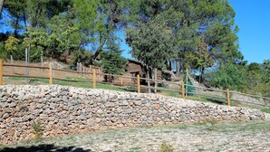 A stone retaining wall with a wooden fence, a wooden structure in the background, and dense trees. - Rural apartment Aptos. Rural Tourism Las Maravillas for 5 people (Jaén)