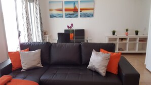 Ein modernes Wohnzimmer mit einem schwarzen Ledersofa, orangefarbenen und weißen Kissen, einem Glastisch und einer weißen Unterhaltungsanlage mit Pflanzen.