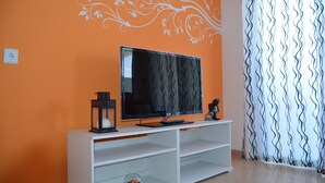Ein Wohnzimmer mit oranger Wand, einem Flachbildfernseher auf einer weißen Unterhaltungsecke, einer kleinen Glasschale für Fische und einem dekorativen Wandbild.