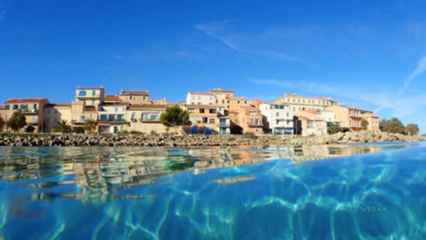A coastal town with buildings along the waterfront, clear blue water, and a rocky shoreline. - L'île-Rousse with its feet in the water (L'Île-Rousse)