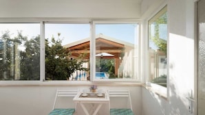 A sunlit room with a white table and two chairs, large windows offering a view of a building with a wooden roof and greenery outside. - Villa Green Oasis by AdriaticLuxuryVillas (Debeljak)