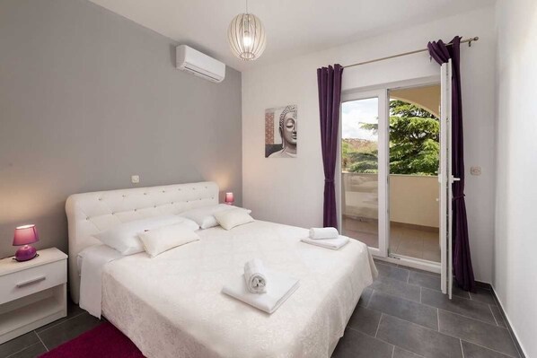 A bedroom with a large bed, a nightstand, a bedside lamp, a window with purple curtains, and a view of greenery outside.