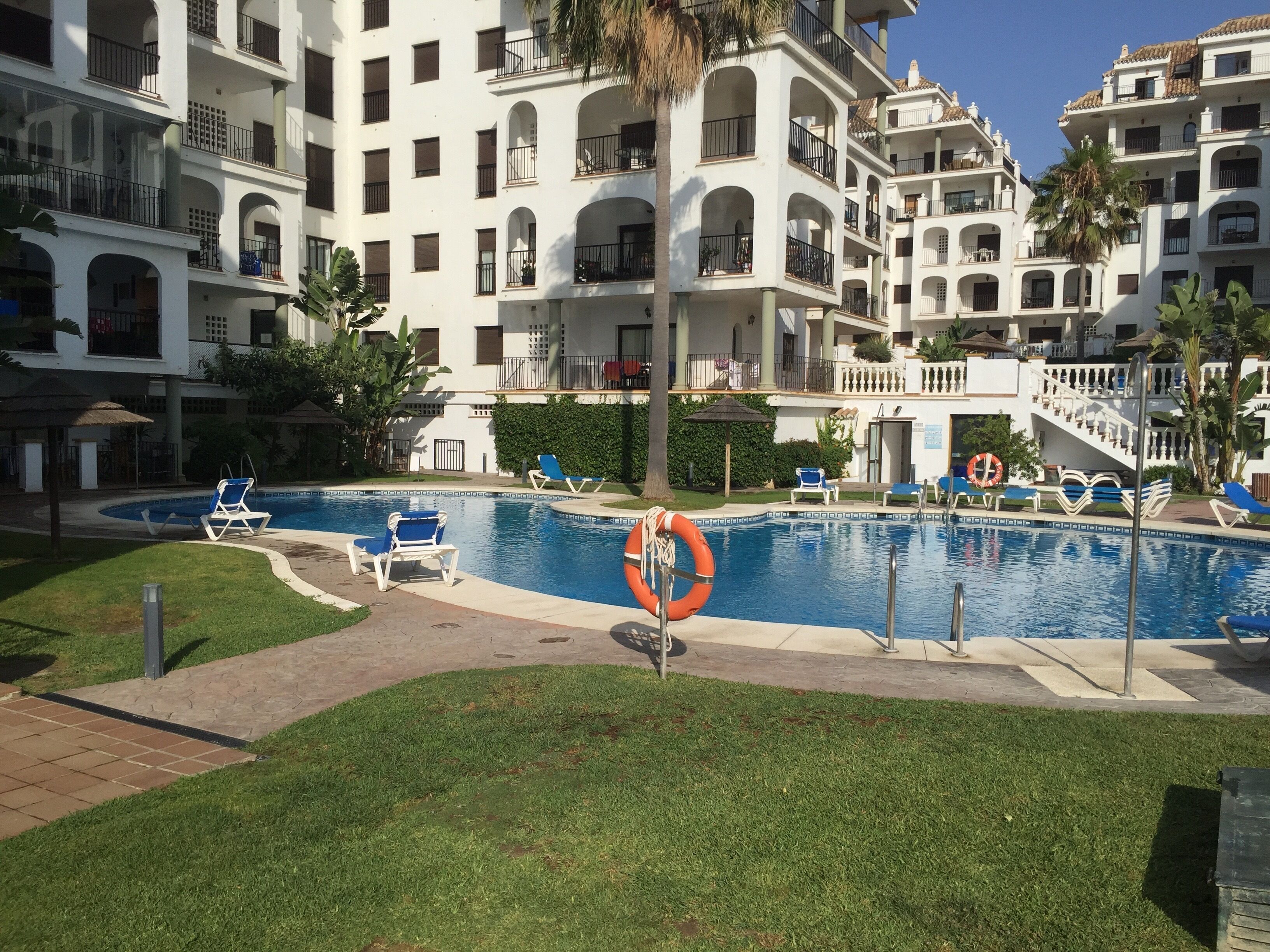 A swimming pool with lounge chairs and a lifebuoy, surrounded by a well-maintained lawn and a multi-story building with balconies.