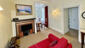 A living room with a fireplace, a red sofa, a dining area, and a hallway leading to a door. - Elegant first floor apartment for 2 people, with great views of mountains.  (Taynuilt)