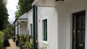 Un bâtiment à l’extérieur blanc, doté de fenêtres à encadrement vert et d’une porte avec une poignée en laiton. On y trouve des espaces extérieurs aménagés avec des meubles en osier et des plantes en pot.