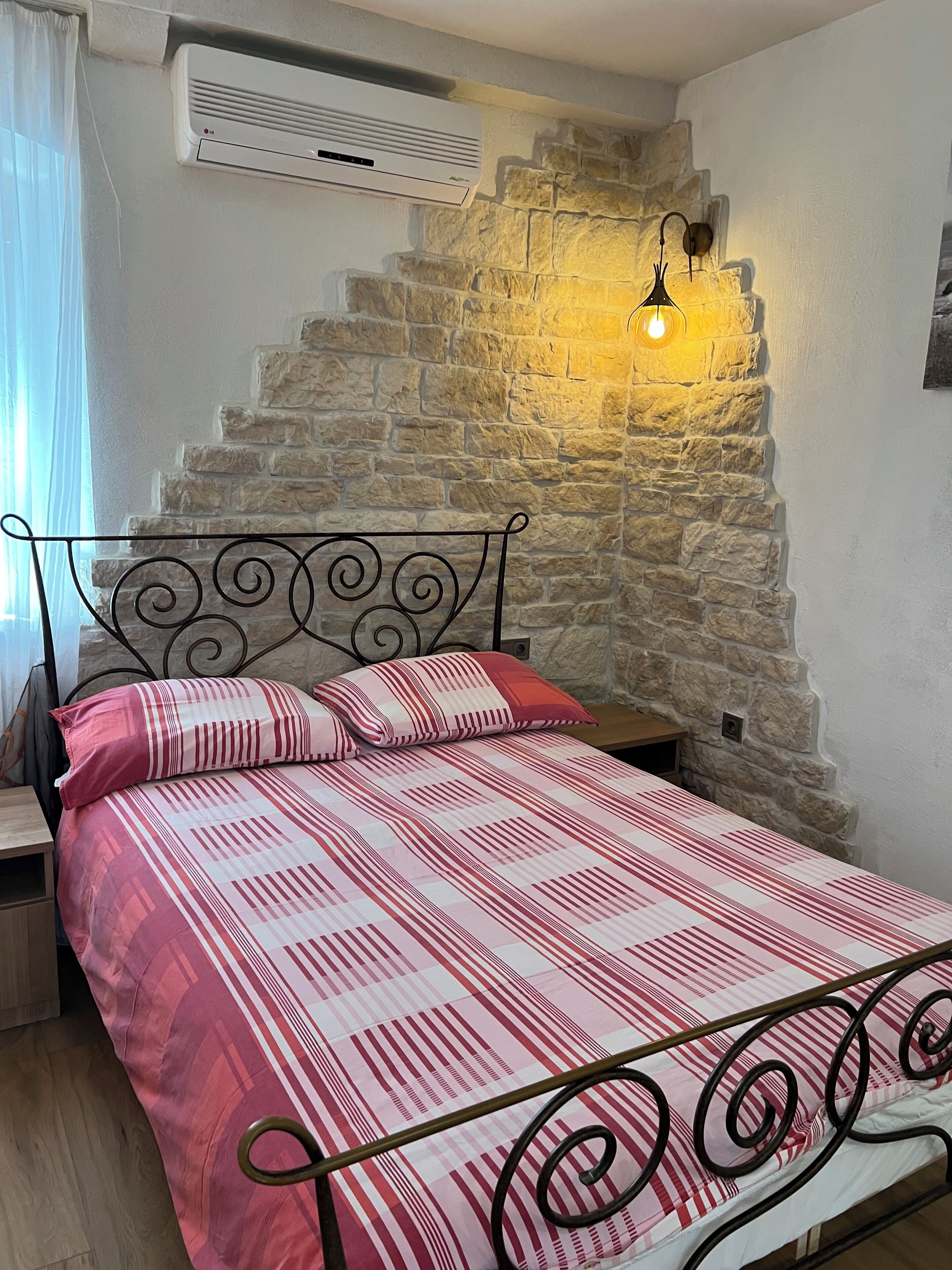 Een slaapkamer met een stenen muur, een bed met metalen frame, een nachtkastje en een wandairconditioner.