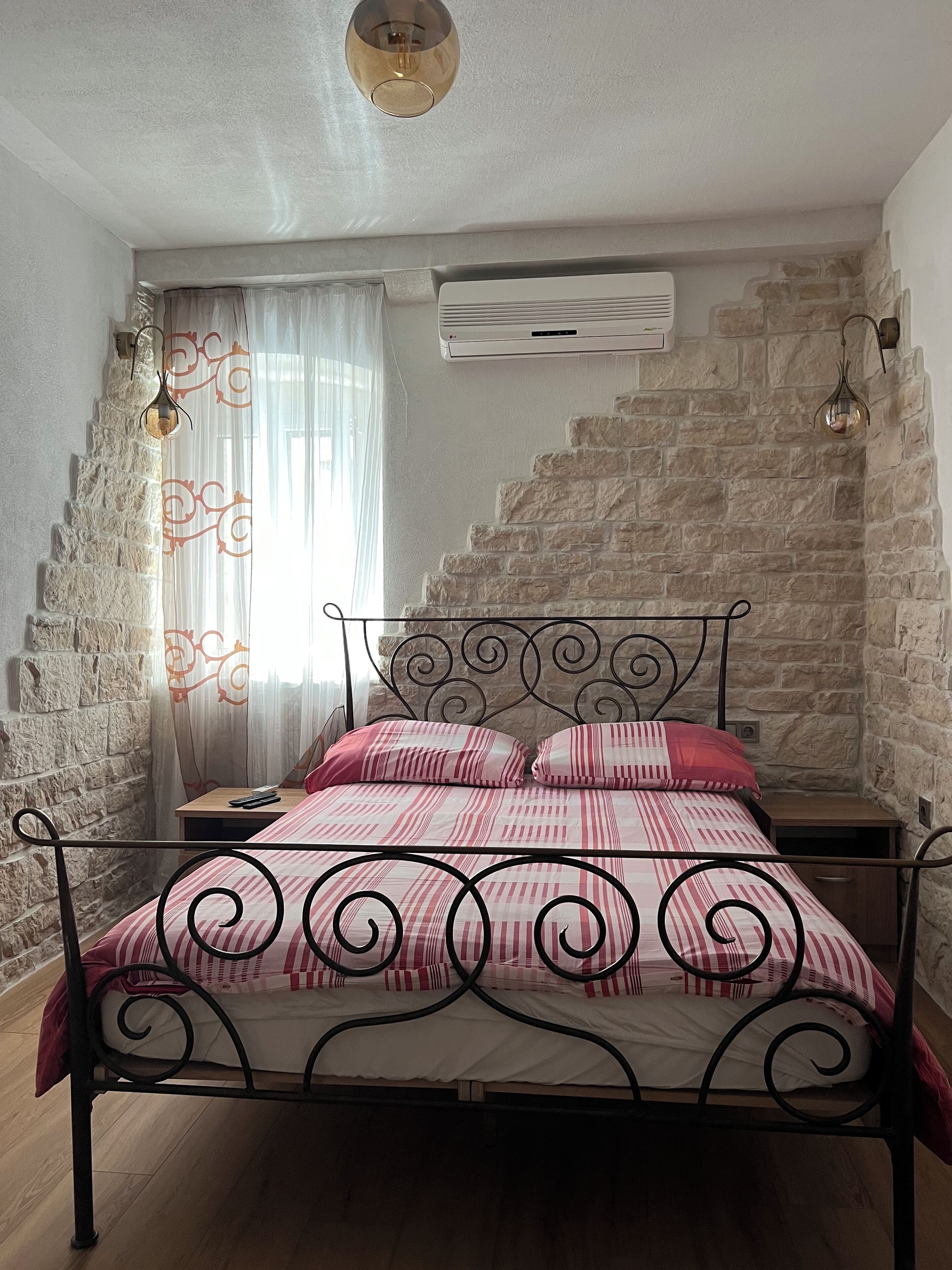 A bedroom with a stone wall, a bed with a metal frame, a nightstand, an air conditioning unit, and a window with curtains.