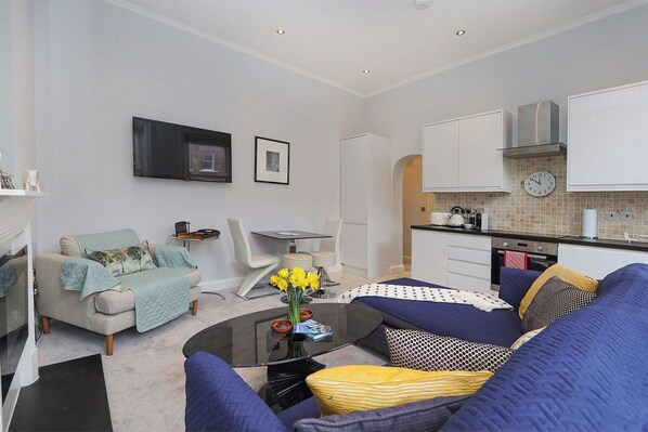 A modern living room with a sofa, coffee table, and a TV. There is a kitchen area with cabinets and a clock on the wall.