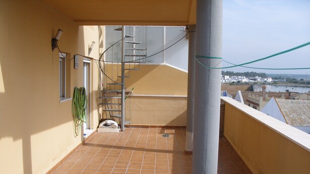 A balcony with a tiled floor, a metal staircase, and a view of a body of water and buildings in the distance. - Magnificent Penthouse in Barbate (Barbate)