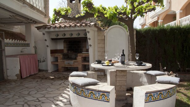 Een terras buiten met een ronde tafel, vier stoelen en een decoratieve, betegelde bank.