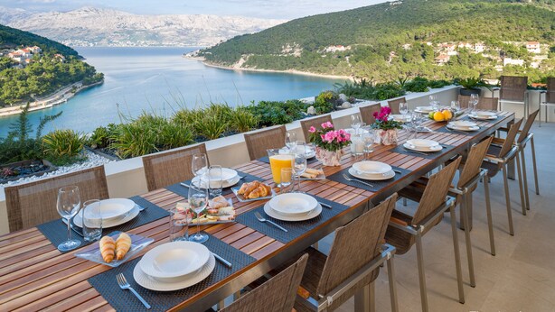 Ein Esstisch gedeckt mit Tellern, Gläsern und Croissants mit Blick auf eine malerische Küstenlandschaft.