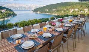 A dining table set with plates, glasses, and croissants, overlooking a scenic coastal view. - Luxurious Seaside Villa. Stunning Views. Private Pool. Spacious Outdoor Living (Pucišca, Brac, Split)