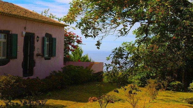 Un edificio rosa con contraventanas verdes, rodeado de un jardín cuidado y árboles.
