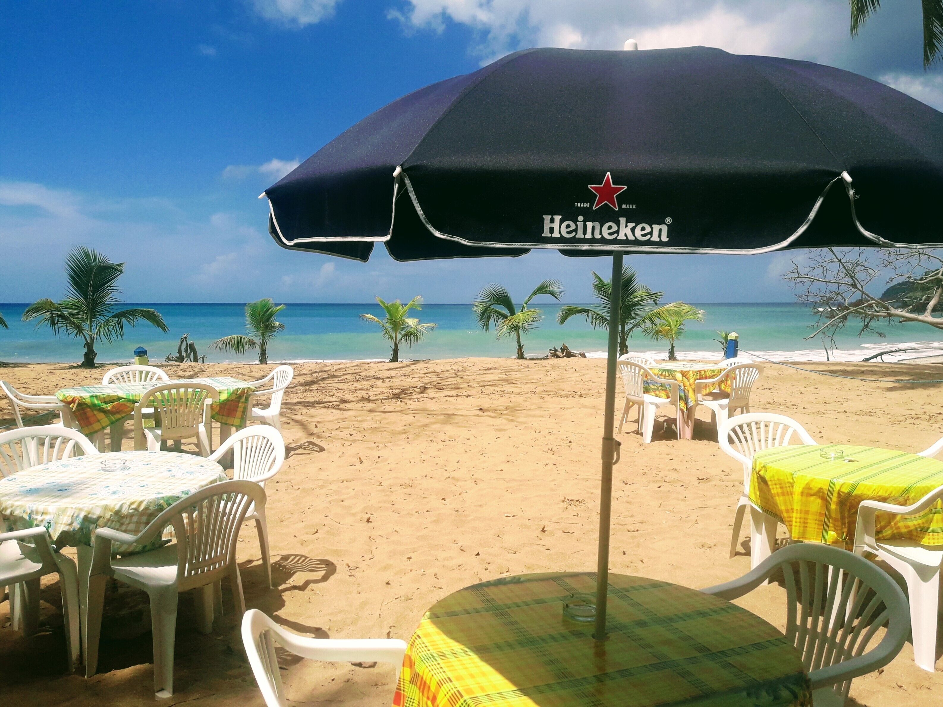 Un espace aménagé avec des tables et des chaises, situé au bord de la plage et abrité par un parasol Heineken, d’où l’on a une vue sur l’océan.