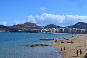 Uma praia com águas azul-claras, afloramentos rochosos e um horizonte urbano ao fundo.