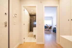 Quarto de hotel moderno com piso de madeira, banheiro com chuveiro de vidro e área de jantar com mesa e cadeiras.