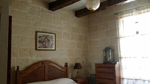A bedroom with a stone wall, a wooden bed, a nightstand with a lamp, a dresser, and a chandelier. - Apartment in the heart of Vittoriosa with AC (il-Birgu)