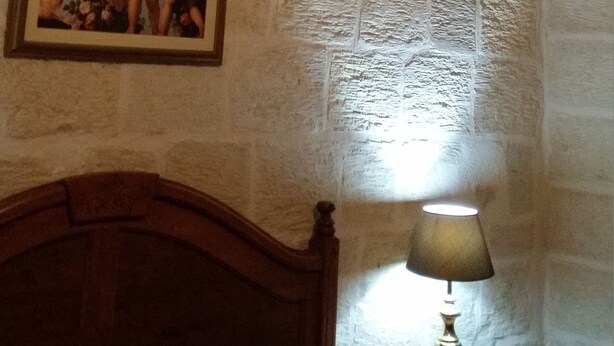 A room with stone walls, a wooden ceiling beam, a bed, a nightstand with a lamp, and a painting on the wall. - Apartment in the heart of Vittoriosa with AC (il-Birgu)