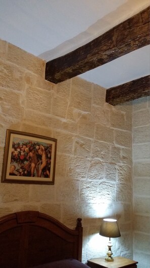 A room with stone walls, a wooden ceiling beam, a bed, a nightstand with a lamp, and a painting on the wall.