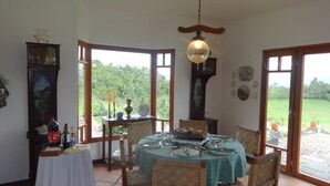 Un comedor con una mesa preparada para cuatro personas, rodeado de sillas de mimbre, un candelabro y vistas a un jardín a través de amplios ventanales.