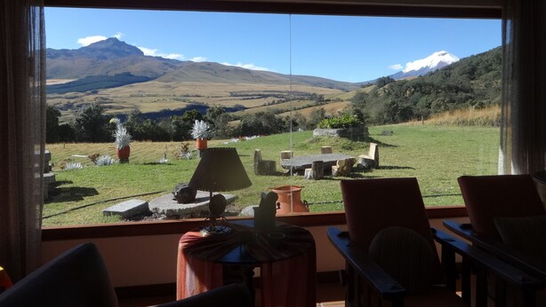 Habitación con un ventanal grande con vistas a un paisaje montañoso. Adentro, hay una mesa con sillas y una lámpara.