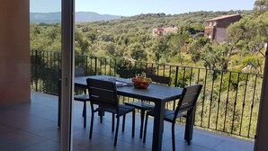 Un balcón con mesa y sillas, un frutero y vistas a un paisaje verde con árboles y edificios.