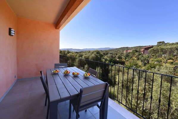 Un balcon avec une table dressée pour le dîner, agrémenté de corbeilles de fruits. Le balcon offre une vue sur un paysage vallonné, avec des arbres et des bâtiments visibles au loin.