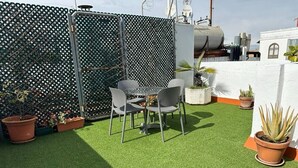 Eine Dachterrasse mit Kunstrasen, einem Metalltisch und -stühlen, umgeben von Pflanzkübeln und einem Zierzaun.