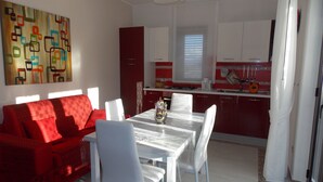 Un salon moderne avec un canapé rouge et des chaises blanches, une table à manger agrémentée d’un vase, et une cuisine avec des meubles rouges et une fenêtre.