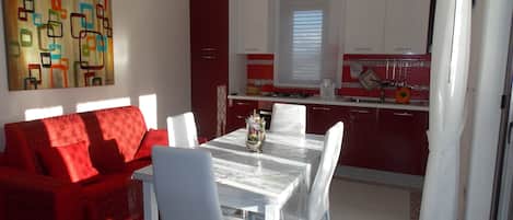 A modern living room with a red sofa and white chairs, a dining table with a vase, and a kitchen with red cabinets and a window.