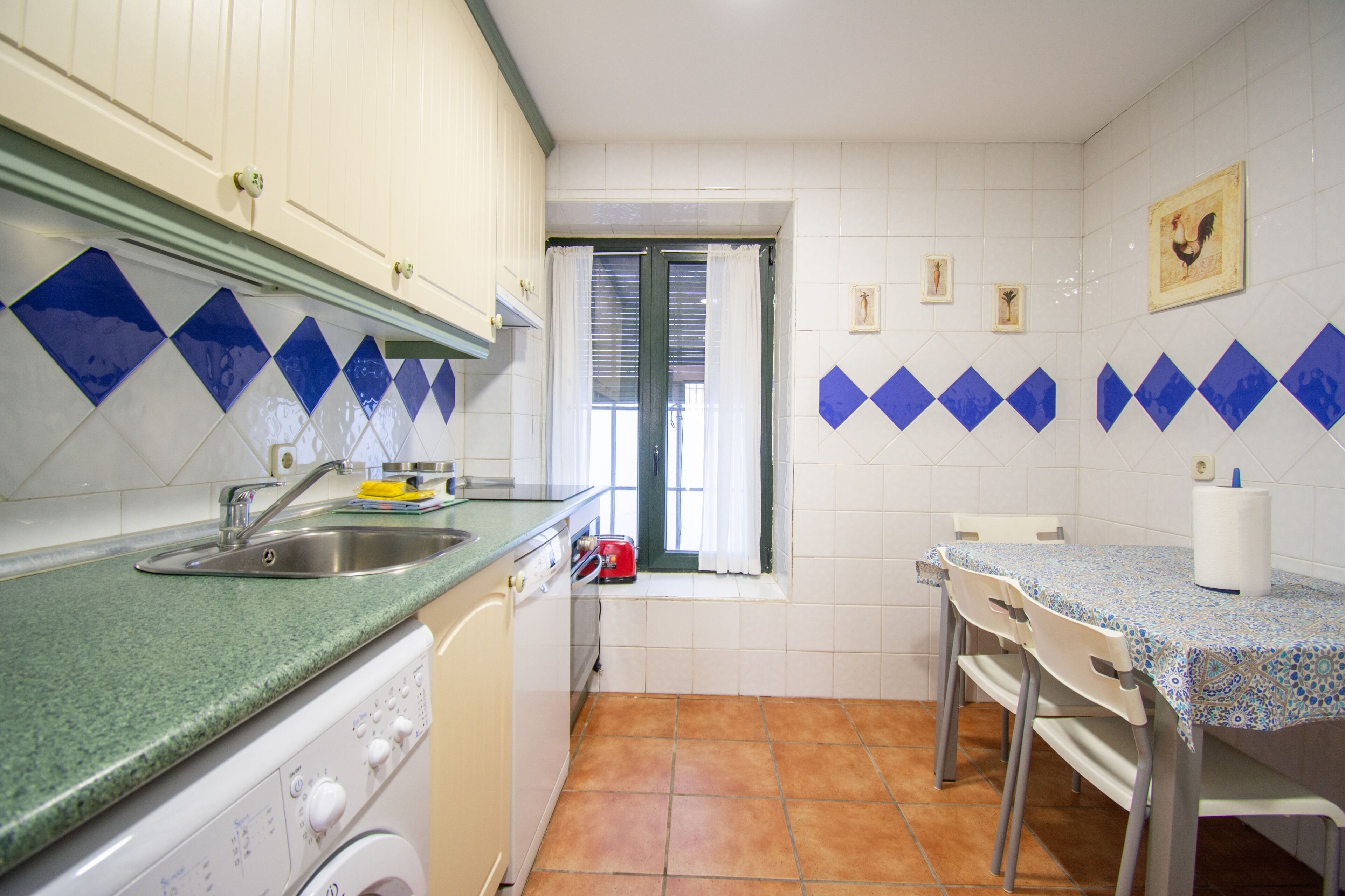 A kitchen with a washing machine, a sink, a window with curtains, and a table with chairs.