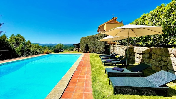 A swimming pool with lounge chairs and umbrellas, surrounded by a stone wall and greenery.