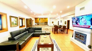 A living room with a black leather sofa, a coffee table, and a TV mounted on the wall. There is a kitchen in the background with wooden cabinets and a dining area with a table and chairs.