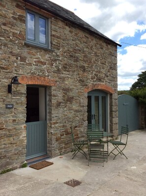 Un bâtiment en pierre avec une porte et des fenêtres, un petit espace extérieur aménagé avec des chaises et une table, et un portail en arrière-plan.