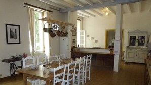 Un coin repas avec une table en bois et des chaises blanches, une cuisine en arrière-plan et une vue sur l’extérieur par une fenêtre.