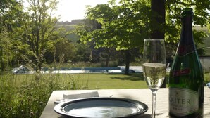 Une bouteille de vin Kriter, un verre et une assiette sur une table en plein air, avec en arrière-plan des arbres et une étendue d’eau.