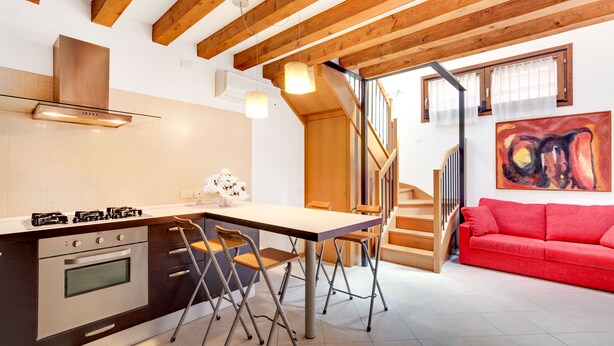 Een moderne keuken met een inbouwoven, een eethoek met een tafel en stoelen, een rode bank en een trap naar een hoger gelegen verdieping.